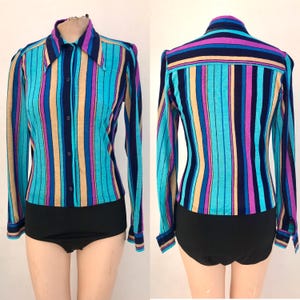 Può includere: Una camicia vintage a maniche lunghe con righe verticali turchesi, blu navy, viola e beige. La camicia ha un colletto e una chiusura con bottoni. La parte inferiore è nera.