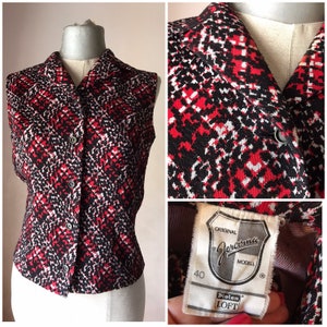 Op de afbeelding: Een mouwloos rood, zwart en wit gepatroneerd knoopshirt met een kraag. Het shirt heeft een label met de tekst "Original Jercoma Modell 40 Diolen Loft".