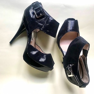 Può includere: Un paio di scarpe nere con tacco alto, punta aperta e cinturino alla caviglia. Le scarpe hanno un dettaglio con fibbia sul cinturino alla caviglia.