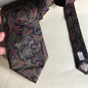 May include: A dark brown paisley patterned tie with a green, brown, and burgundy color scheme. The tie is folded and laid out on a light beige surface.