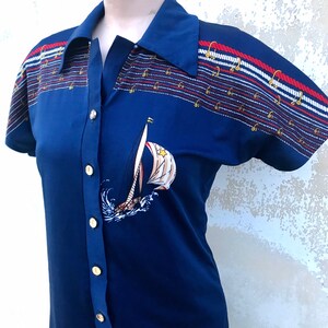 May include: A blue short-sleeved dress with a white sailboat graphic and red, white and blue striped trim. The dress has a collared neckline and gold buttons down the front.