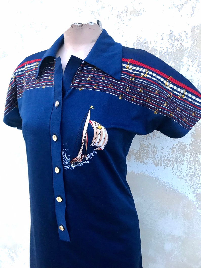 May include: A blue short-sleeved dress with a white sailboat graphic and red, white and blue striped trim. The dress has a collared neckline and gold buttons down the front.