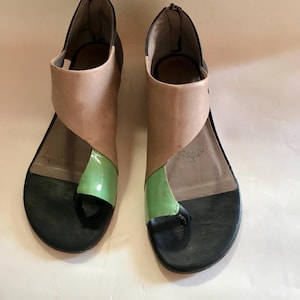 Op de afbeelding: Een paar sandalen met open teen met een uniek ontwerp. De sandalen hebben zwarte zolen, lichtbruine en groene accenten en een ritssluiting bij de hiel. De sandalen hebben een modern, asymmetrisch ontwerp.