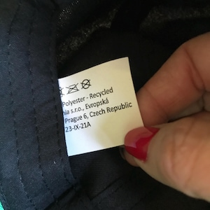 Op de afbeelding: Wit etiket met zwarte tekst die "Polyester - Gerecycled hia s.r.o., Evropsk&aacute; Praag 6, Tsjechische Republiek 23-IX-21A" luidt.