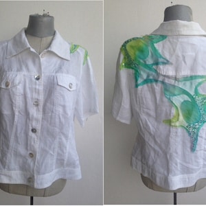 以下が含まれることがあります： 白いリネン素材のボタンダウンシャツ。半袖で、背面に緑と青のフローラルデザインが施されています。
