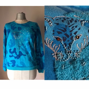 May include: A blue sweatshirt with a camouflage pattern and a beaded embroidered image of a leopard. The leopard is in the center of the sweatshirt and is surrounded by a blue camouflage pattern.