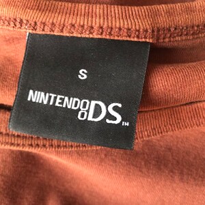May include: A black tag with white text that reads "Nintendo DS" and "S" in the top right corner. The tag is attached to a brown shirt.