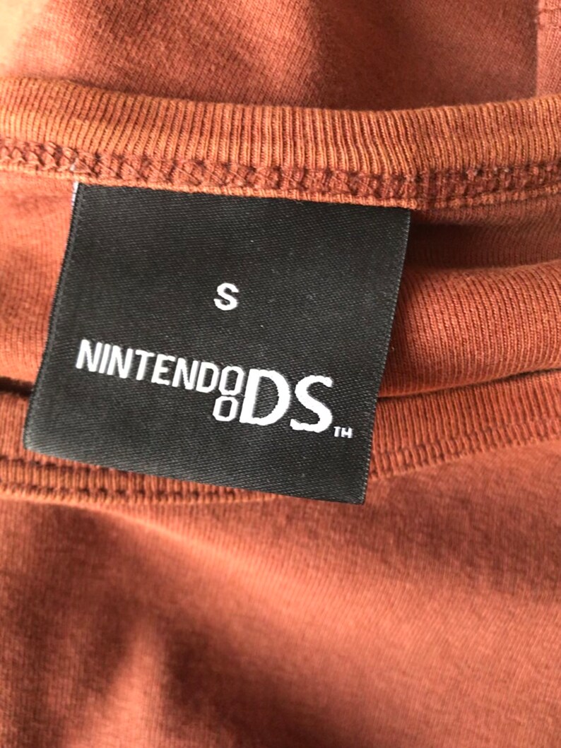 May include: A black tag with white text that reads "Nintendo DS" and "S" in the top right corner. The tag is attached to a brown shirt.