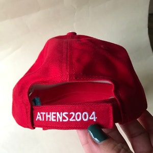 Puede incluir: Gorra de b&eacute;isbol roja con bordado blanco que dice "ATHENS 2004".
