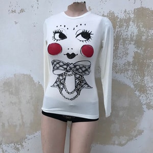 May include: White long-sleeved shirt with a black and white illustration of a face with red cheeks, a bow, and a necklace.