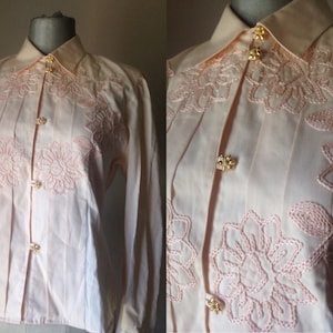 May include: A pale pink long-sleeved blouse with a button-up front and delicate floral embroidery. The blouse has a classic collar and gold-toned buttons.