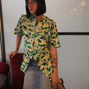 May include: A woman wearing a green, yellow and blue patterned button-down shirt with a denim skirt. She has pink hair and is wearing glasses.