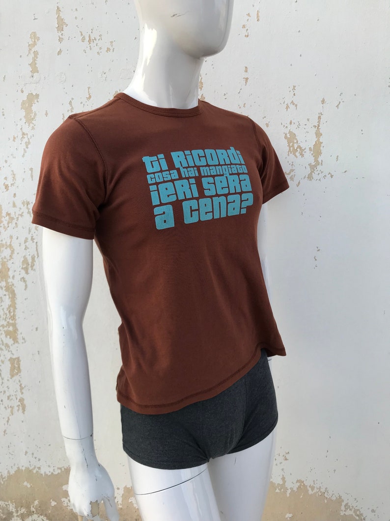 May include: A brown t-shirt with a blue graphic that says "Ti Ricordi cosa hai mangiato ieri sera a cena?"
