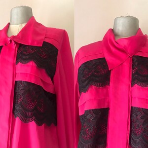 Può includere: Una blusa rosa a maniche lunghe con bordino in pizzo nero. La blusa ha un colletto e una chiusura con bottoni sul davanti.