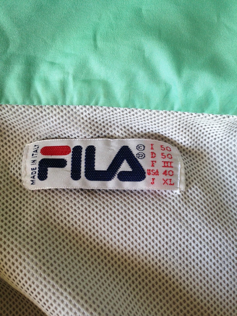 Puede incluir: Primer plano de una etiqueta Fila, con el nombre de la marca en azul marino, rojo y blanco. La etiqueta est&aacute; cosida sobre una tela de malla beige claro, con una tela verde menta encima. La etiqueta indica que la prenda fue fabricada en Italia.