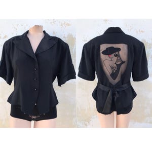 以下が含まれることがあります： 後ろに透けるパネルがあり、帽子をかぶった女性と背景にエッフェル塔が刺繍された、黒いボタン留めの半袖ブラウスです。