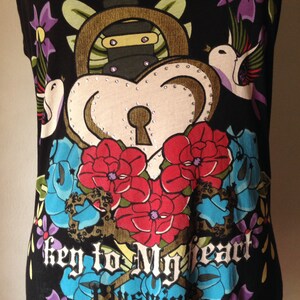 Op de afbeelding: Zwart tanktop met een kleurrijke grafische print van een hart met een sleutelgat, bloemen en vogels. De tekst "Key to My Heart" is gedrukt op de voorkant van het shirt.