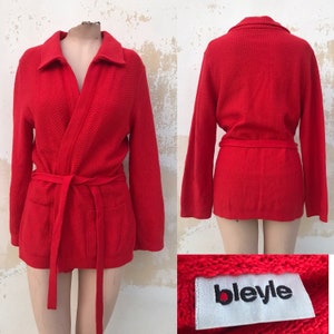 Könnte beinhalten: Rote Strickjacke im Wickelstil mit einem selbstbindenden Gürtel. Die Jacke hat eine Waffelstrickstruktur und ein Etikett mit der Aufschrift "bleyle".