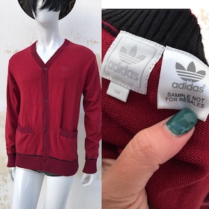 May include: Red cardigan sweater with a v-neck, button closure, and two pockets. The sweater has black trim around the neckline, cuffs, and hem. The Adidas logo is embroidered on the left chest. The tag reads "Adidas Sample Not For Resales".