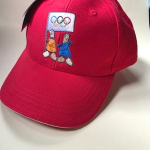 Puede incluir: Gorra de b&eacute;isbol roja con un dise&ntilde;o bordado de dos figuras sosteniendo una bandera con los anillos ol&iacute;mpicos. Las figuras visten ropa naranja y azul.