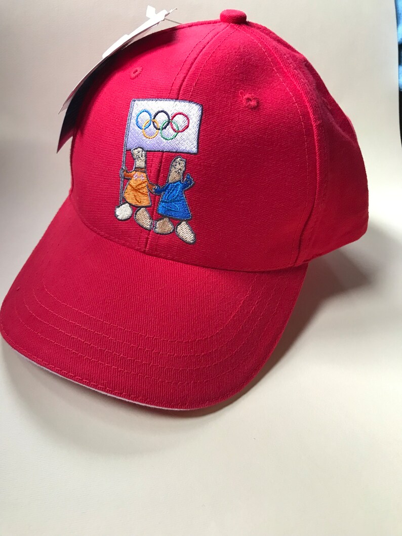Puede incluir: Gorra de b&eacute;isbol roja con un dise&ntilde;o bordado de dos figuras sosteniendo una bandera con los anillos ol&iacute;mpicos. Las figuras visten ropa naranja y azul.