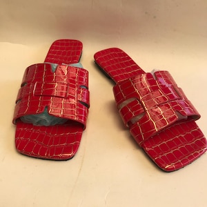May include: A pair of red sandals with a glossy, crocodile-effect texture. The sandals have a square toe and multiple straps across the top. The soles are black.
