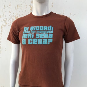 May include: A brown t-shirt with a blue graphic print that says "Ti Ricordi cosa hai mangiato ieri sera a cena?"