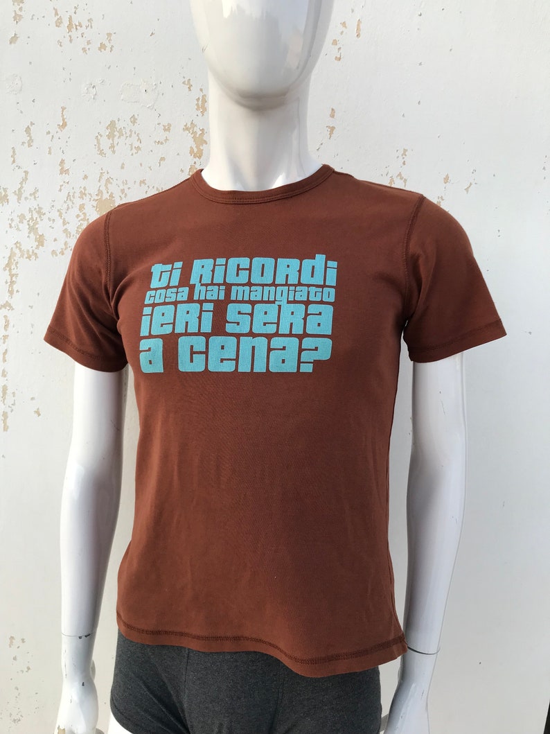 May include: A brown t-shirt with a blue graphic print that says "Ti Ricordi cosa hai mangiato ieri sera a cena?"