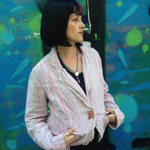 May include: A woman with black hair and pink highlights is wearing a pink and white striped blazer with a ruffled trim. She is standing in front of a blue and green graffiti wall. The text on the wall reads "Give your time or your money".