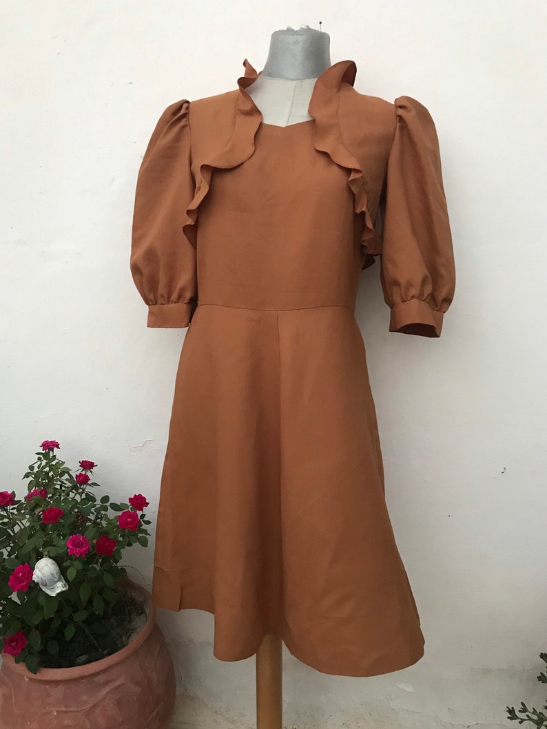 May include: A brown vintage dress with a ruffled neckline and puffed sleeves. The dress has a fitted bodice and a flowing skirt.