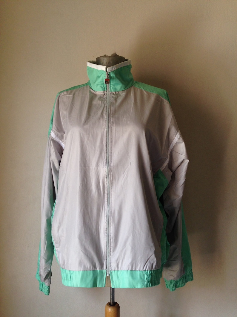 Puede incluir: Chaqueta deportiva vintage gris y verde menta con cierre de cremallera completo. La chaqueta presenta un cuello alto con ribete blanco y detalles en verde menta en los hombros y el dobladillo. La chaqueta se exhibe en un maniqu&iacute;.