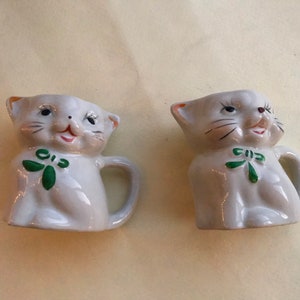 May include: Two white ceramic cat figurines with green bows. The cats are sitting upright with their heads tilted and their mouths open in a smiling expression.