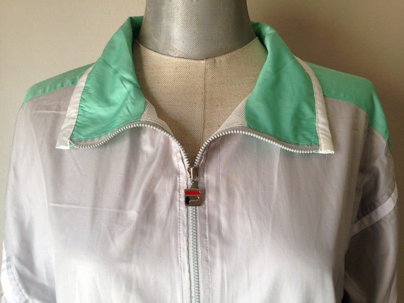 Puede incluir: Chaqueta deportiva Fila vintage en gris claro con detalles en verde menta y blanco. La chaqueta tiene una cremallera completa y un peque&ntilde;o logotipo de Fila en el tirador. El cuello y los hombros son de color verde menta.