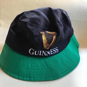 Op de afbeelding: Een zwarte en groene bucket hat met het Guinness-logo en een gouden harp met de tekst "ESTD 1759".