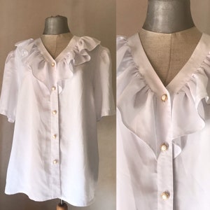 以下が含まれることがあります： フリル襟と半袖の白いボタンダウンブラウス。ブラウスは白の中央に金色のボタンが付いています。