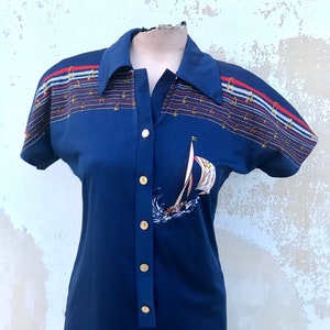 May include: A navy blue short-sleeved dress with a white sailboat graphic and gold buttons. The dress has a striped pattern with red, white, and blue accents.