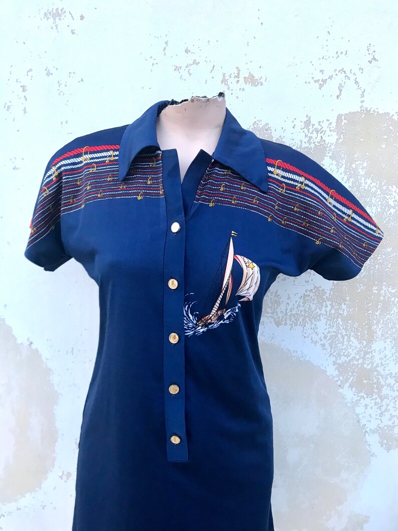 May include: A navy blue short-sleeved dress with a white sailboat graphic and gold buttons. The dress has a striped pattern with red, white, and blue accents.