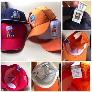 Puede incluir: Tres gorras de b&eacute;isbol, una roja, una naranja y una azul. La gorra roja tiene los anillos ol&iacute;mpicos y una figura de dibujos animados. La gorra naranja tiene los anillos ol&iacute;mpicos y la palabra "Atenas" en ella. La gorra azul tiene una figura de dibujos animados.