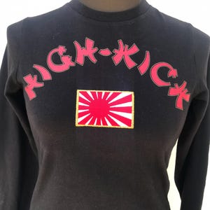 Può includere: Maglietta nera a maniche lunghe con la scritta rossa "HIGH-KICK" ad arco sul petto. Sotto, un disegno a bandiera rossa e bianca con bordo dorato. La maglietta è esposta su un manichino.
