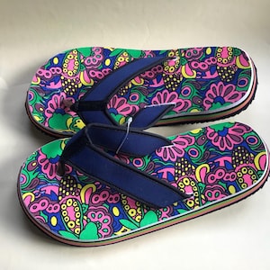 May include: A pair of colorful flip-flops with a vibrant floral pattern in pink, green, yellow, and blue. The flip-flops have navy blue straps and a white and black border.