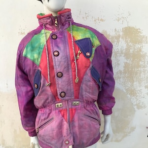 Può includere: Una giacca da sci vintage con un motivo tie-dye in tonalità rosa, viola, verde e blu. La giacca ha un colletto alto, una chiusura con cerniera e diverse tasche.
