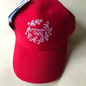 Puede incluir: Gorra de b&eacute;isbol roja con un dise&ntilde;o bordado en blanco que presenta los anillos ol&iacute;mpicos y el texto "ATHENS 2004".