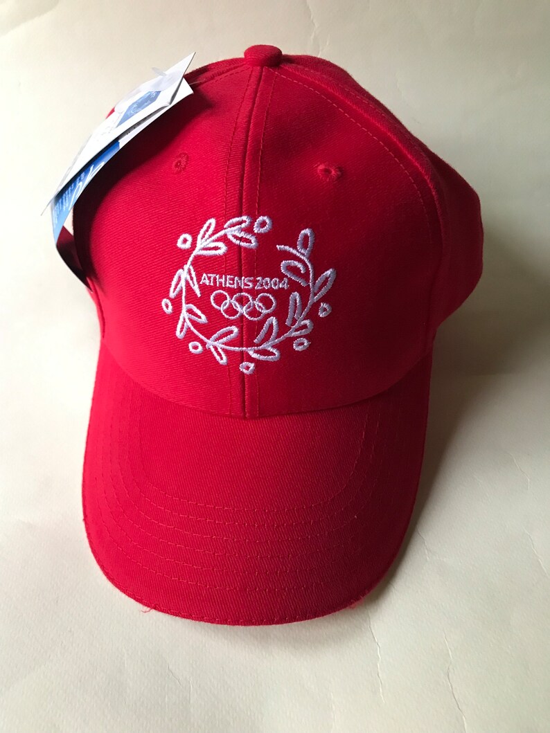 Puede incluir: Gorra de b&eacute;isbol roja con un dise&ntilde;o bordado en blanco que presenta los anillos ol&iacute;mpicos y el texto "ATHENS 2004".