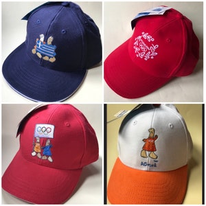 Puede incluir: Cuatro gorras de b&eacute;isbol con diferentes dise&ntilde;os. La primera gorra es azul marino con costuras blancas y un dise&ntilde;o bordado blanco de dos ping&uuml;inos sosteniendo una bandera con los anillos ol&iacute;mpicos. La segunda gorra es roja con costuras blancas y un dise&ntilde;o bordado blanco de una corona con los anillos ol&iacute;mpicos. La tercera gorra es roja con costuras blancas y un dise&ntilde;o bordado blanco de dos ping&uuml;inos sosteniendo una bandera con los anillos ol&iacute;mpicos. La cuarta gorra es blanca con costuras naranjas y un dise&ntilde;o bordado blanco de un ping&uuml;ino sosteniendo una bandera con el texto "Atenas 2004".