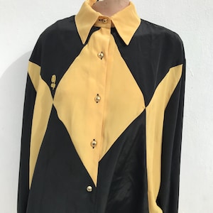以下が含まれることがあります： 印象的な幾何学模様の長袖ブラウス。 黒と黄色のカラーブロックパターンで、黄色の襟と金色のボタンが付いています。 左側には小さな花のエンブレムが見えます。 ブラウスはシルクのような素材で作られています。