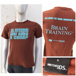 May include: A brown t-shirt with a blue graphic that says "ti Ricordi cosa hai mangiato ieri Sera a cena?" on the front and "allena La tua mente co BRAIN TRAINING&trade; SU NINTENDEDS." on the back. The shirt also has a small black tag that says "S NINTENDODS.&trade;".