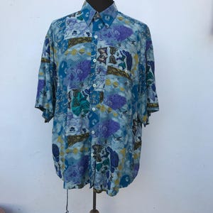 May include: A short-sleeved button-down shirt with a vibrant, all-over pattern. The shirt features a mix of blue, purple, and yellow geometric and floral designs. It has a classic collar and is displayed on a mannequin.