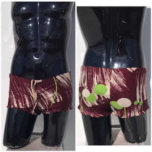 May include: Burgundy and cream swim shorts with a drawstring waist. The shorts feature a pattern of cream brushstrokes and green and cream floral accents. The shorts are displayed on a black mannequin.
