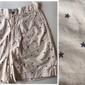 以下が含まれることがあります： 黒の星柄のベージュのショートパンツ。ショートパンツはボタンとジッパーで留め、前面にポケットが2つ付いています。