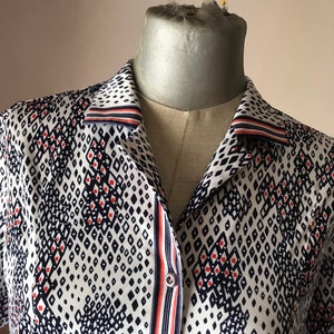 Könnte beinhalten: Ein weißes Hemd mit einem geometrischen Muster aus marineblauen Rauten und kleinen roten Punkten. Der Kragen und die Knopfleiste weisen Streifen in Marineblau, Weiß und Orange auf. Das Hemd wird auf einer Schaufensterpuppe präsentiert.